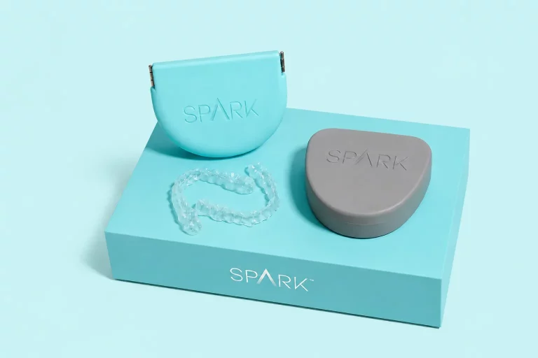 Transparenter SPARK-Zahnaligner und zwei Aufbewahrungsboxen auf einer helltürkisen Box vor einem helltürkisen Studiohintergrund.